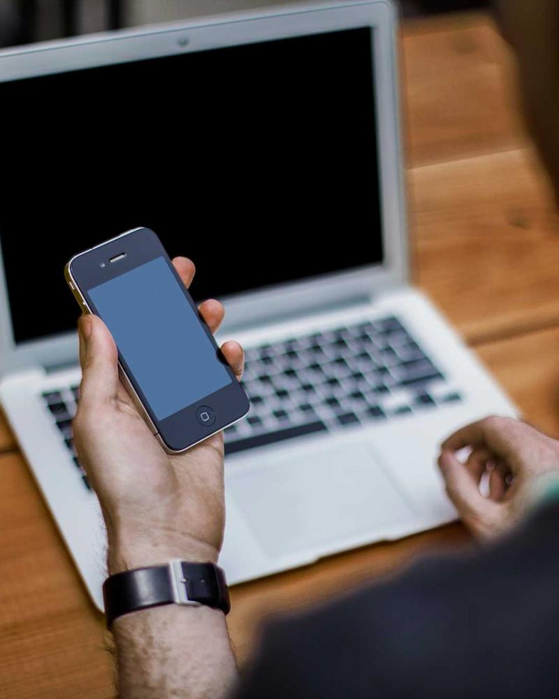 A hand holding a smartphone next to a laptop on a wooden table